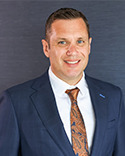 Justin Hofford, a man in a dark blue suit jacket and a patterned brown tie.